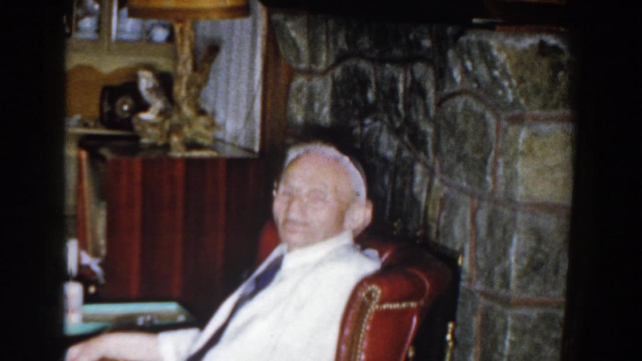 Elderly man smiling in a red armchair with stone fireplace and mantel behind him.