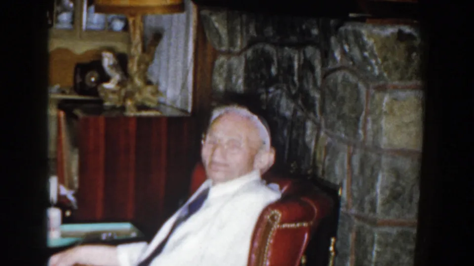 The seated elderly man smiles toward the camera in a 1959 Michigan home interior with a stone fireplace and mantel decor.