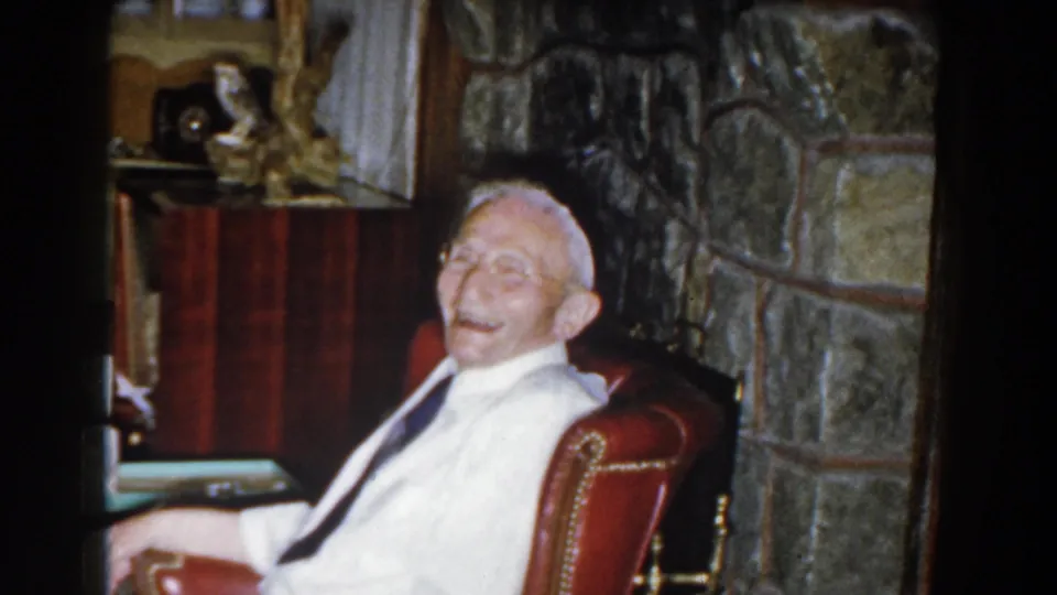An elderly man laughs while seated in a red leather chair in a Michigan living room filmed on 8mm in 1959.