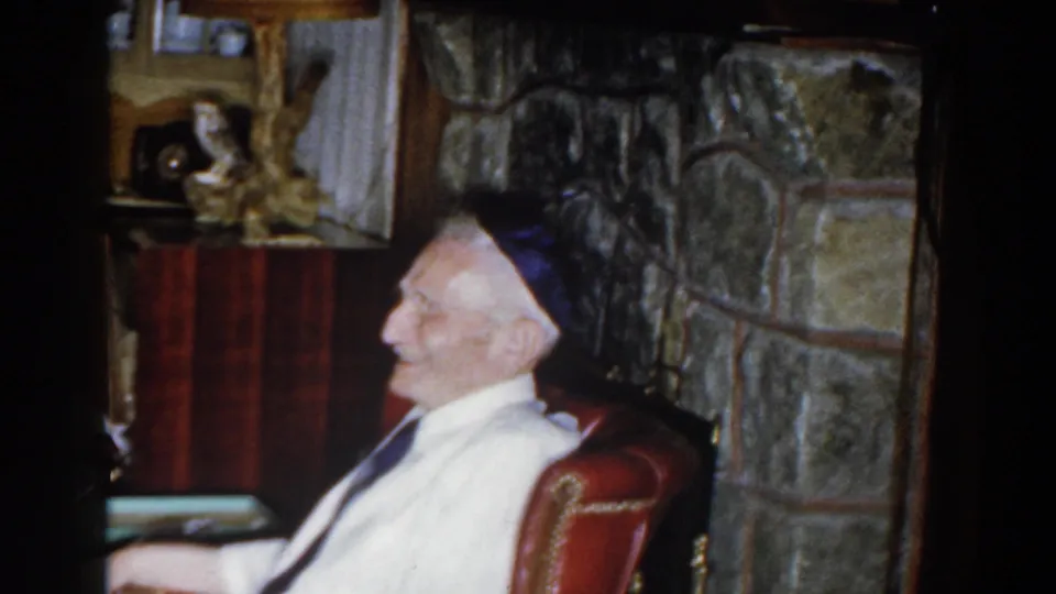 The elderly man turns in profile while seated in the red armchair, with the stone fireplace wall visible behind him.