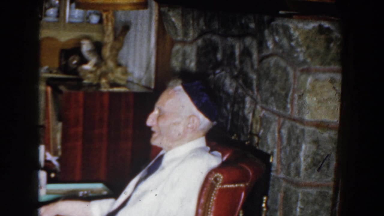 Elderly man in an armchair beside a stone fireplace in a living room.