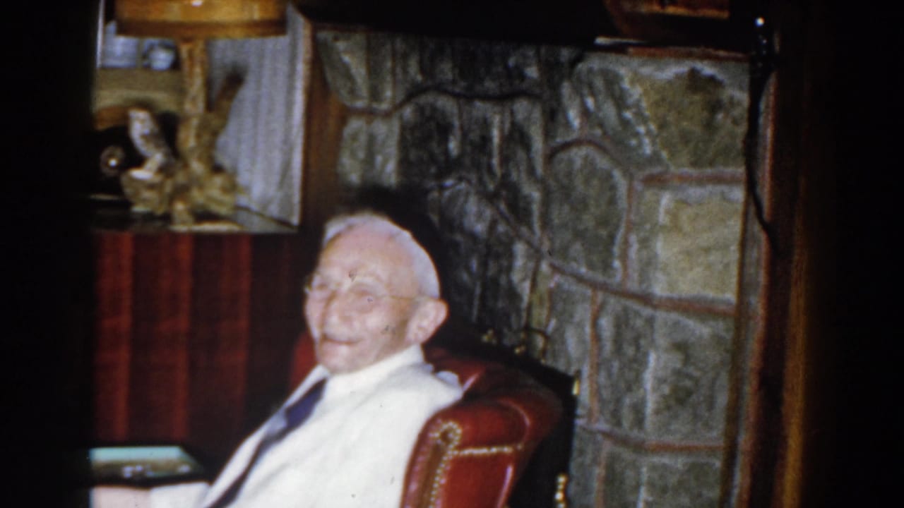 Smiling elderly man seated in a red armchair near a stone fireplace.