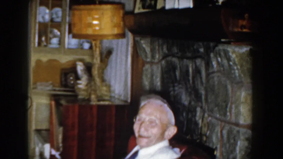 A soft 8mm frame shows the man low in the frame with the stone fireplace, wood cabinet, lamp, and household objects behind him.