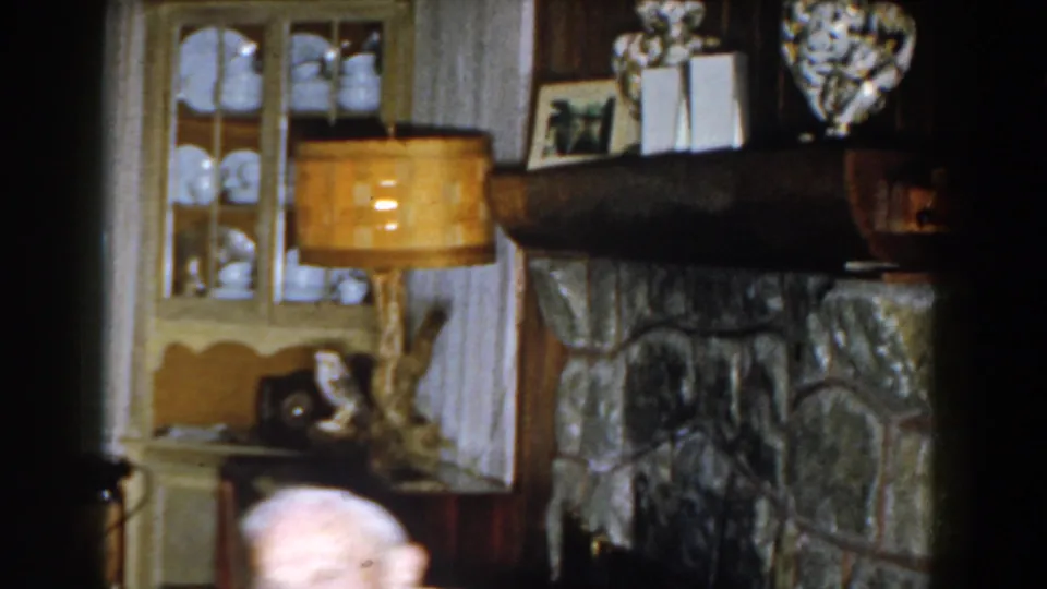 The frame tilts across the 1959 Michigan living room, showing the fireplace, lamp, cabinet, and the seated man's head near the lower edge.