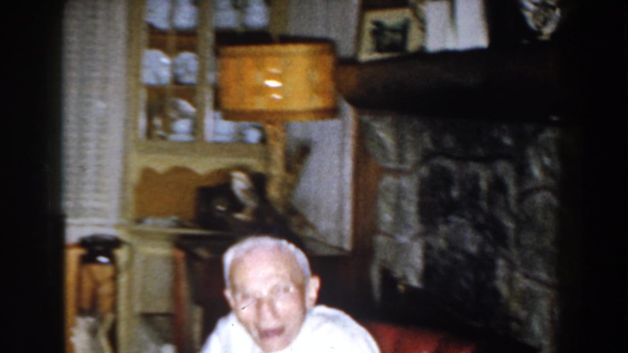 Elderly man seated low in a red armchair beside a fireplace and lamp.