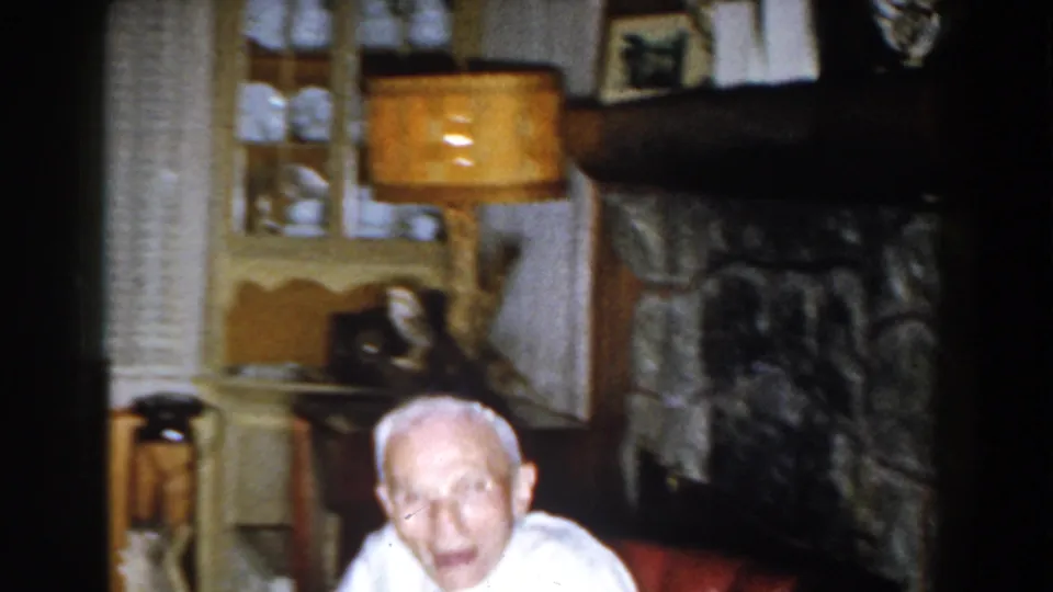 In a 1959 Michigan home-movie frame, an elderly man sits in a red armchair near a stone fireplace, lamp, cabinet, and window.
