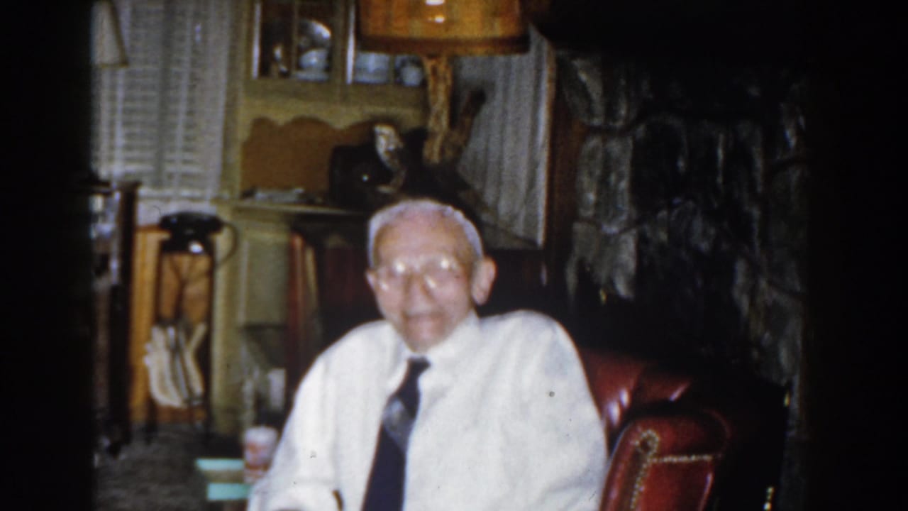 Elderly man in white shirt and tie sitting in a red armchair indoors.