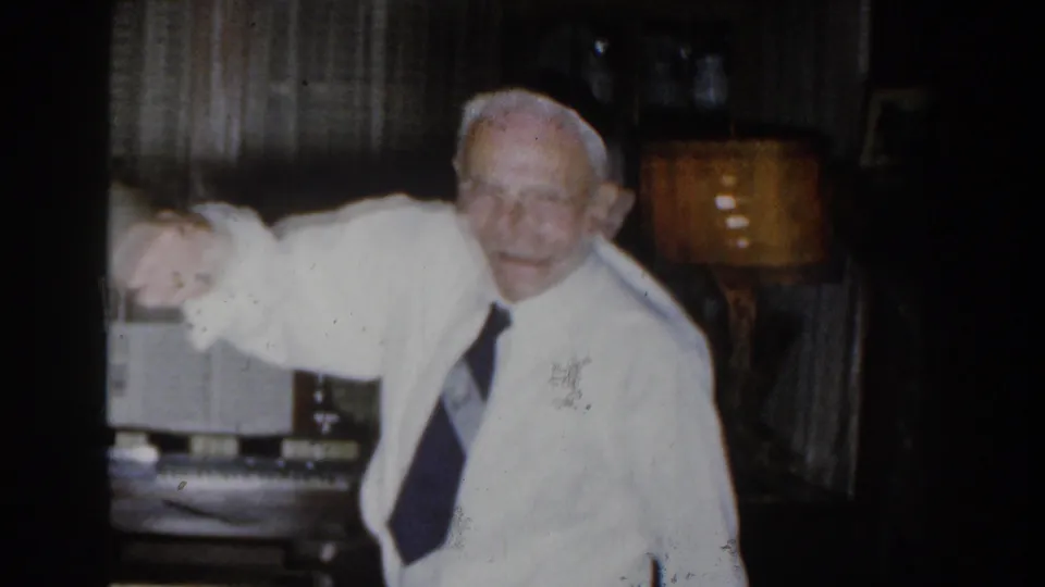 The man begins to stand or gesture from the red chair, his white shirt and tie bright against the darker living room background.