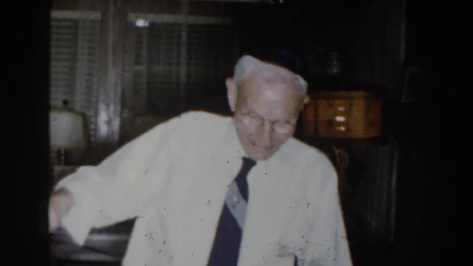 Near the end of the 8mm clip, the man leans forward from the red armchair, looking down as the lamp and window remain visible.