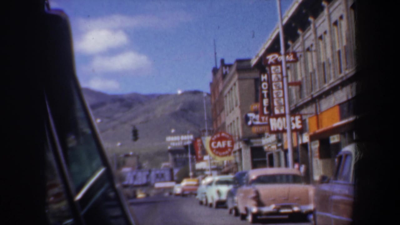 Car-window view down a 1950s street with parked cars, storefront signs, traffic lights, and hills beyond.