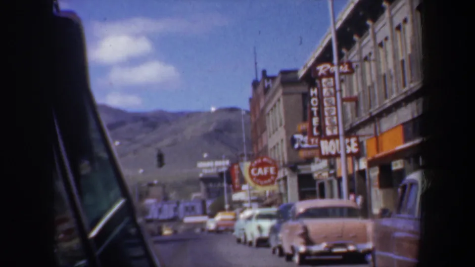 1957 Oregon car-window view of a main street with classic parked cars, hotel and cafe signs, traffic lights, and mountains in the distance.