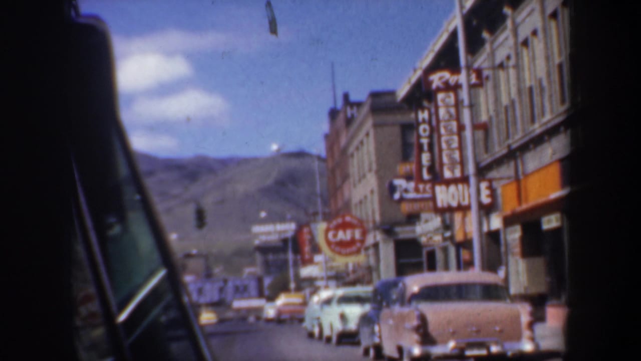 Street view from inside a car with parked classic cars, hotel signage, cafe sign, storefronts, and hills.