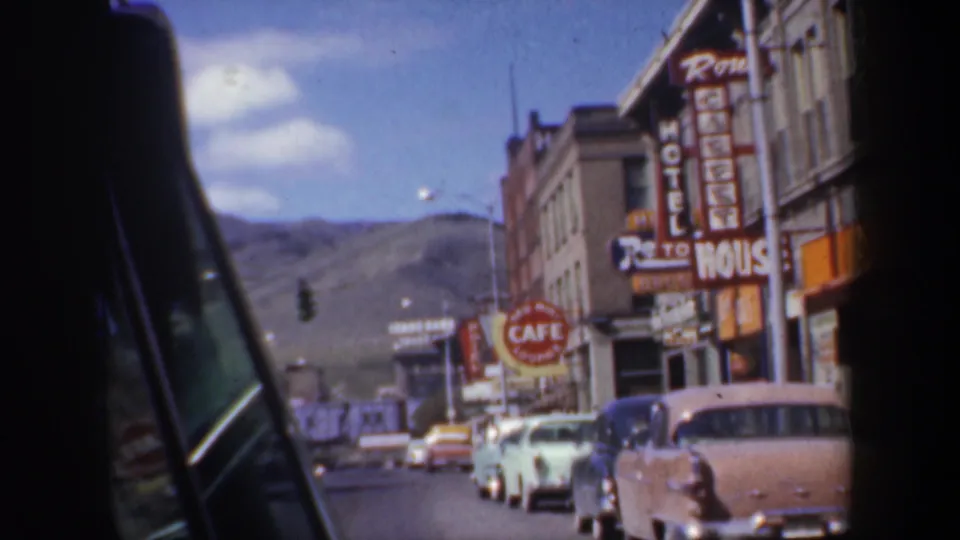 Passenger-window view in 1957 Oregon showing a storefront row, cafe and hotel signs, parked classic cars, and a mountain backdrop.