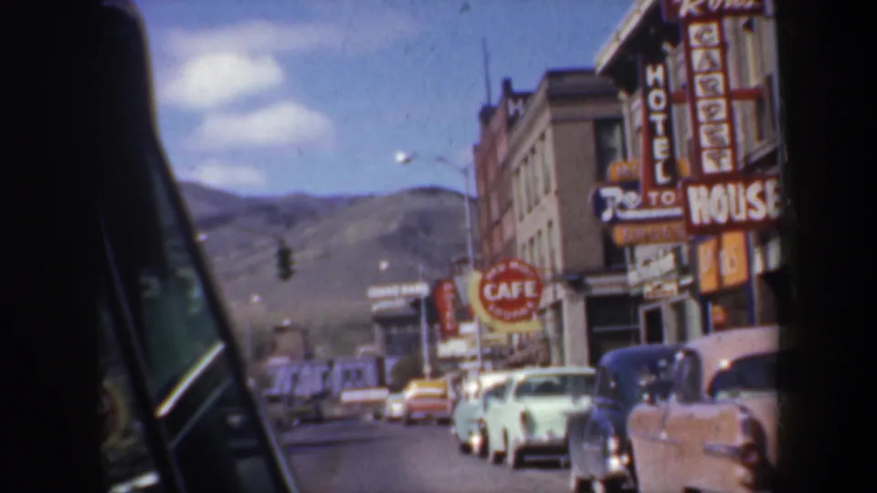 1957 Oregon main street view from a car, showing carpet, hotel, and cafe signs above parked classic cars with hills in the distance.