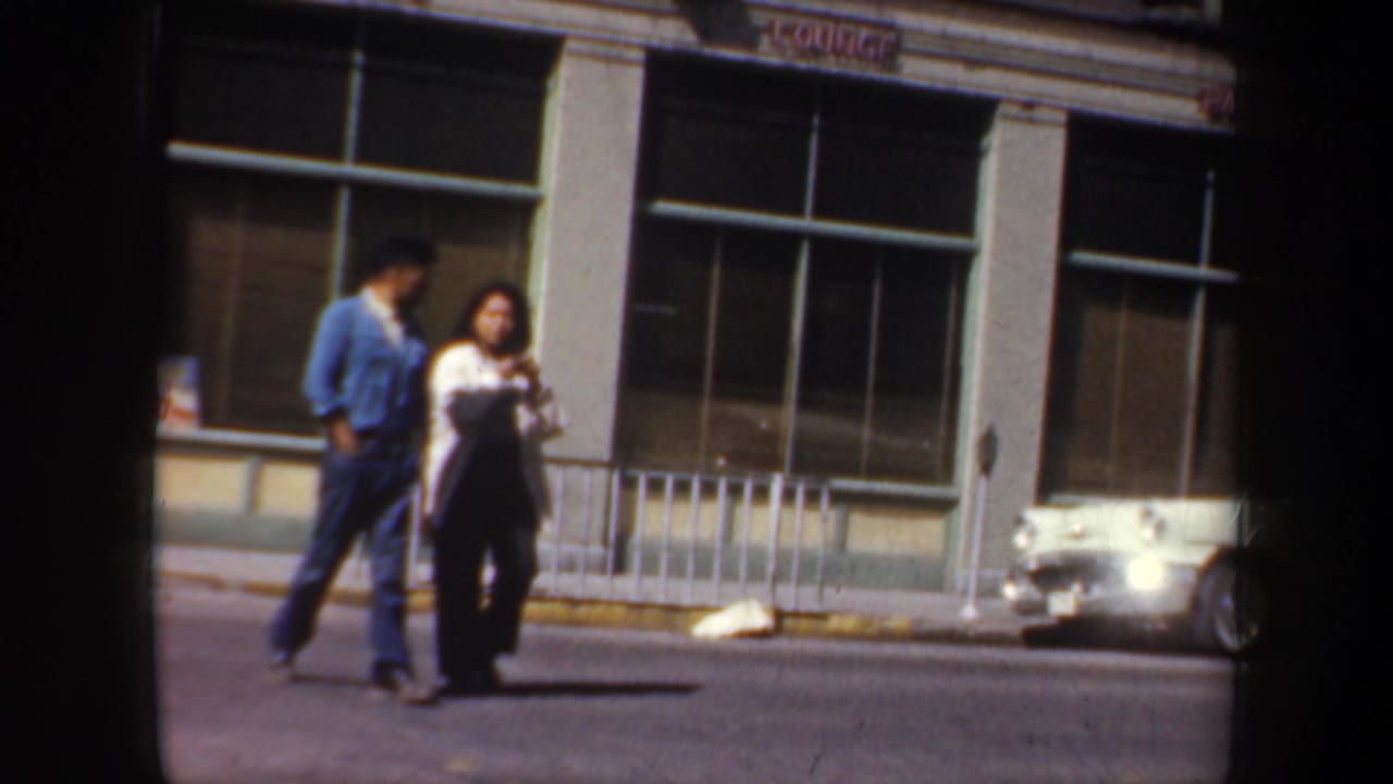 1957 Oregon home-movie frame of an adult couple crossing a sunlit street near storefront windows, parked cars, and a LOUNGE sign.