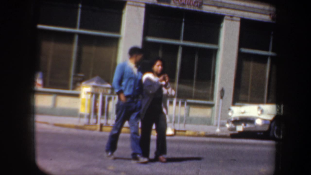 Adult couple walking across the street with storefront windows and parked cars behind them.