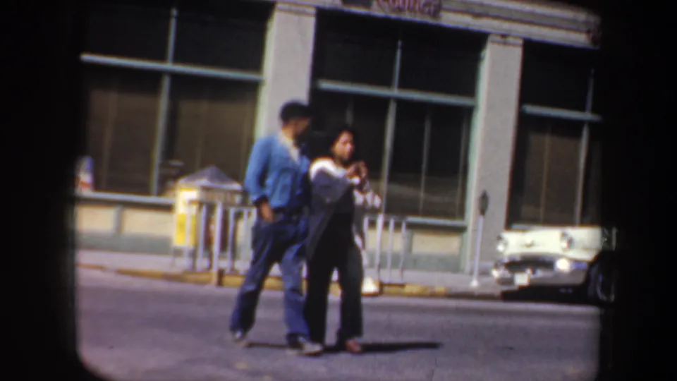 1957 Oregon street scene showing an adult couple mid-crossing, with a storefront, railing, and parked cars in the background.