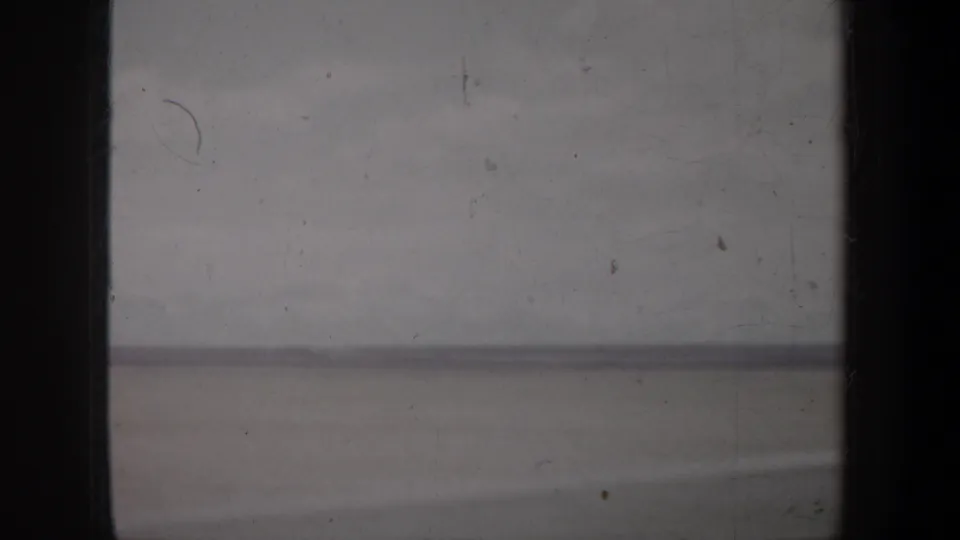 A scratched 1957 Oregon home-movie frame shows a low horizon and open, pale terrain with softened color and focus.