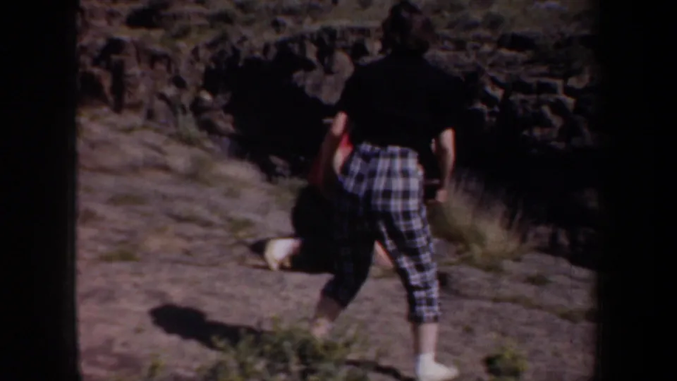 Two women in casual 1950s travel clothing walk away along a rocky Oregon trail bordered by dry brush and stone.