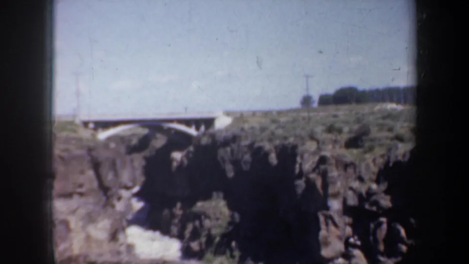 Final Oregon 8mm frame showing the arched bridge, rocky creek channel, utility poles, and sunlit landscape.