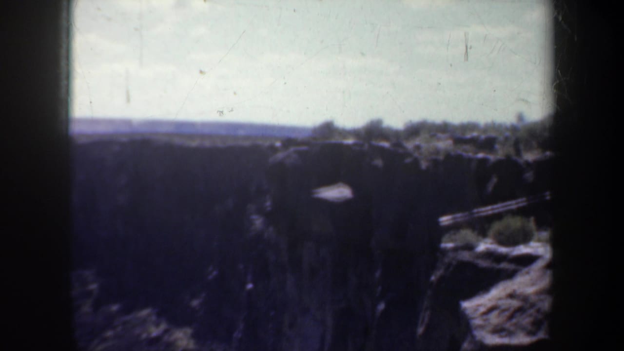 Dark canyon landscape with rocky ledges, scrub vegetation, bright sky, and distant horizon.