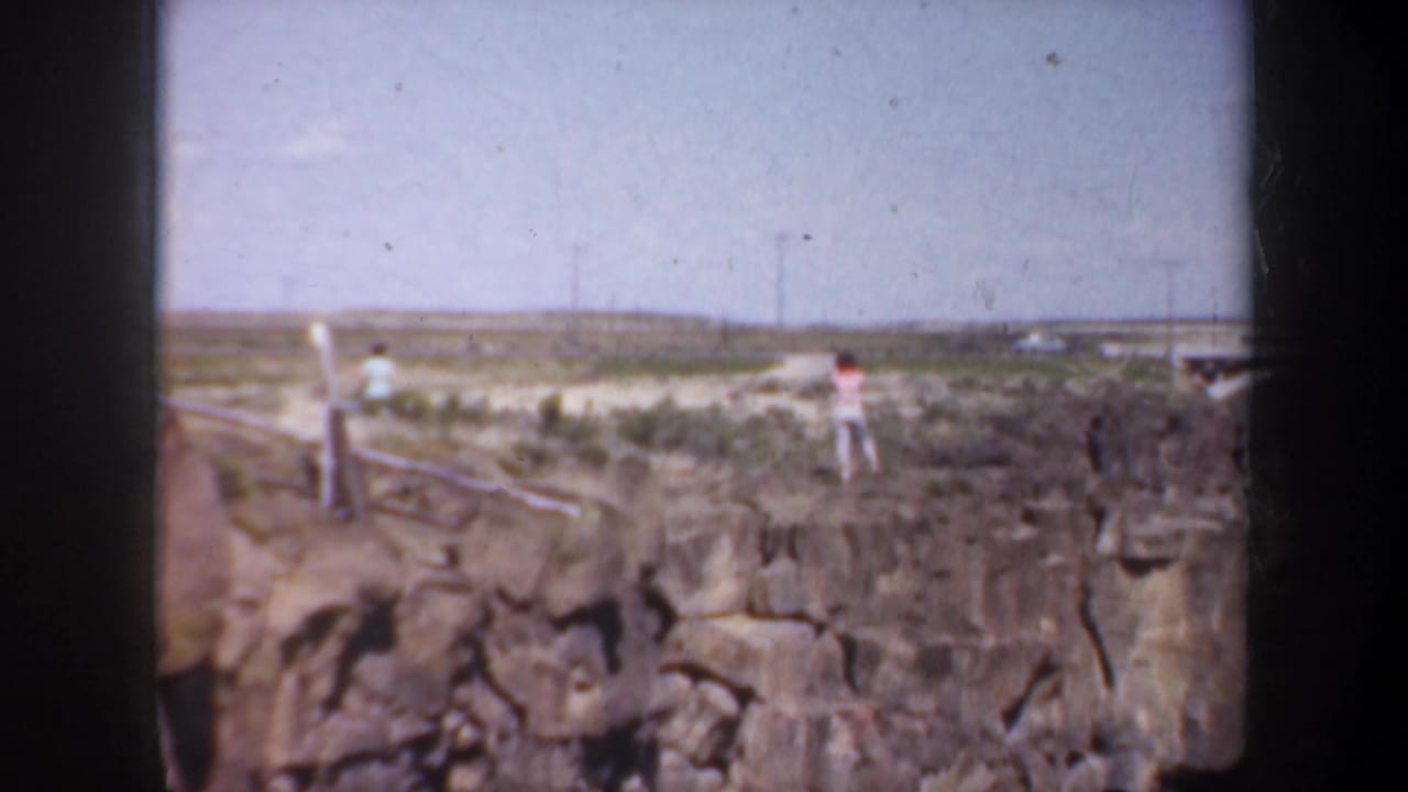 Woman and another distant figure stand on a rocky canyon edge under a pale sky.