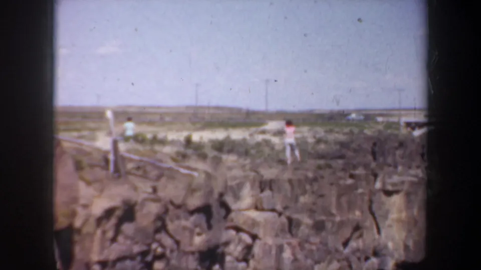 1957 Oregon home-movie frame showing visitors on a rocky canyon rim with scrubland, power lines, and distant infrastructure.