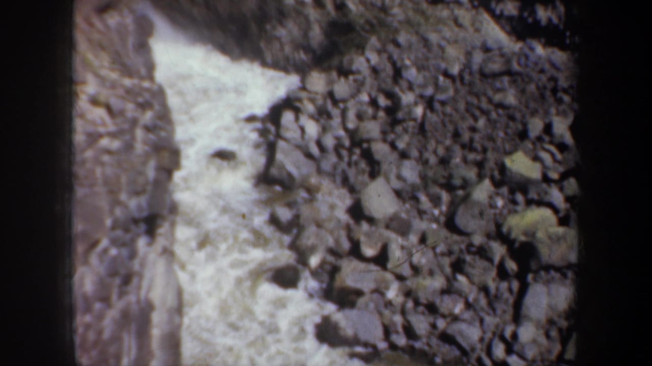 1957 Oregon home-movie frame of foaming river water beside a field of grey boulders and canyon rock.