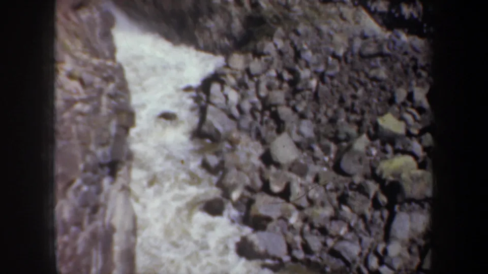1957 Oregon home-movie frame of foaming river water beside a field of grey boulders and canyon rock.