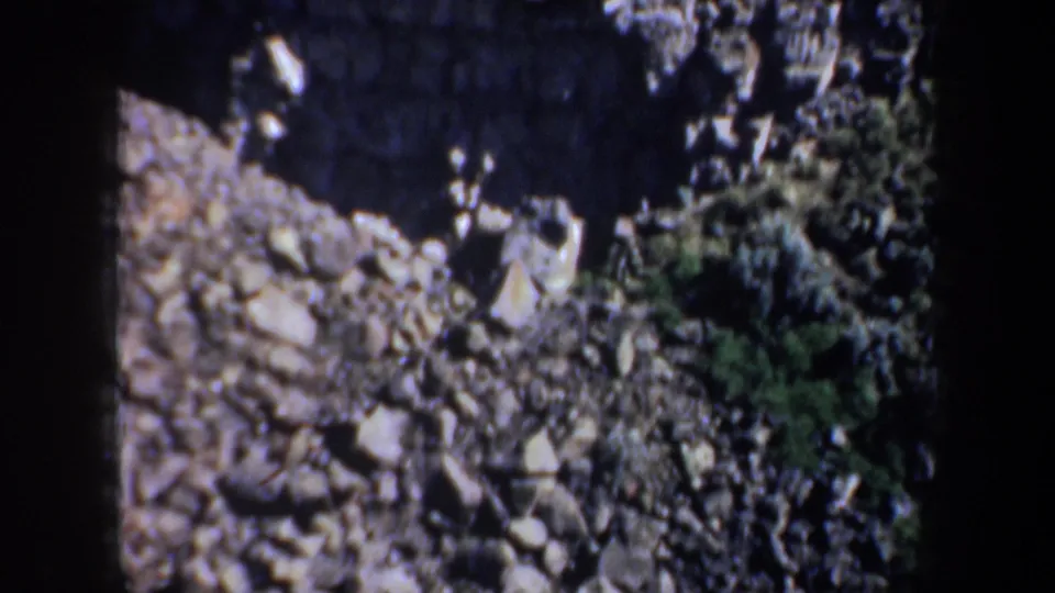1957 Oregon 8mm frame looking toward a rocky slope below dark cliff shadows, with scattered stones and sparse green vegetation.
