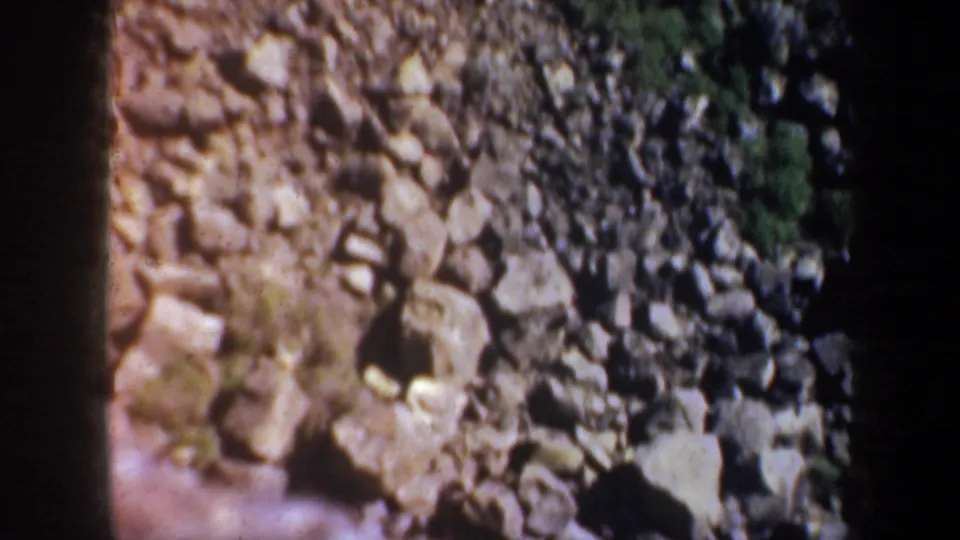 1957 Oregon 8mm frame showing a boulder-strewn riverbank above pale moving water, with dark film-edge borders.