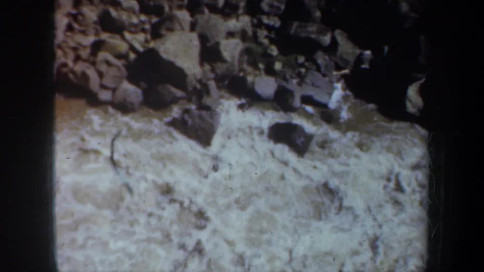 1957 Oregon 8mm frame showing pale rushing river water beneath a boulder-strewn bank, with heavy shadow along the right edge.