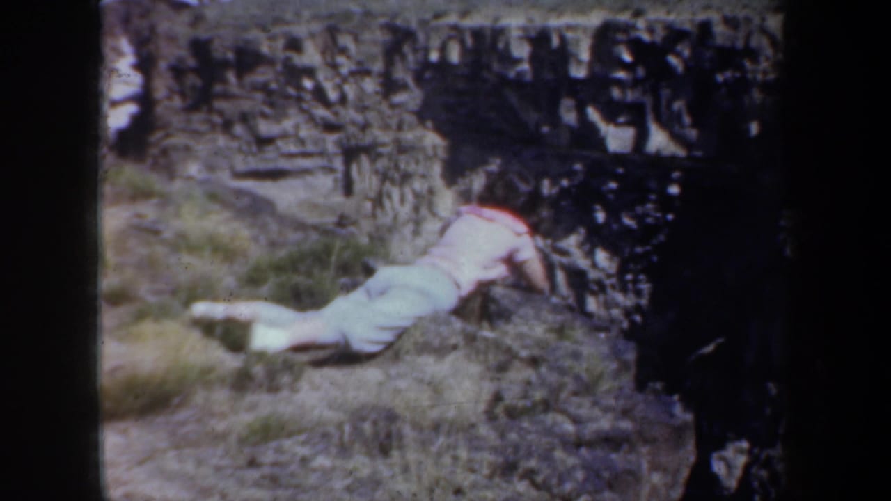1957 Oregon 8mm frame showing a person in light clothing on the ground beside rough stonework in a rural setting.