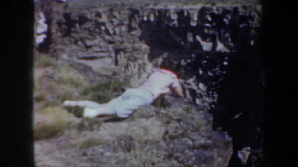 1957 Oregon 8mm frame showing a person in light clothing on the ground beside rough stonework in a rural setting.