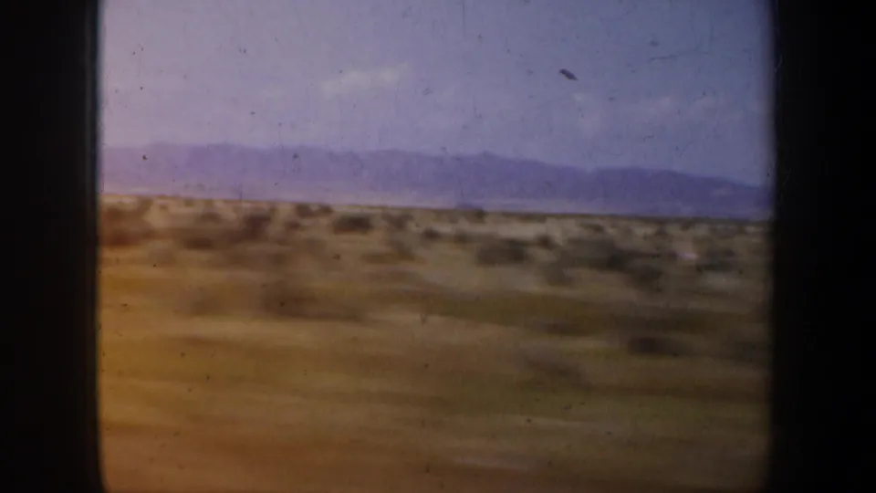 1957 Oregon 8mm frame of dry brown rural terrain with scattered brush and distant mountains under a blue-violet sky.