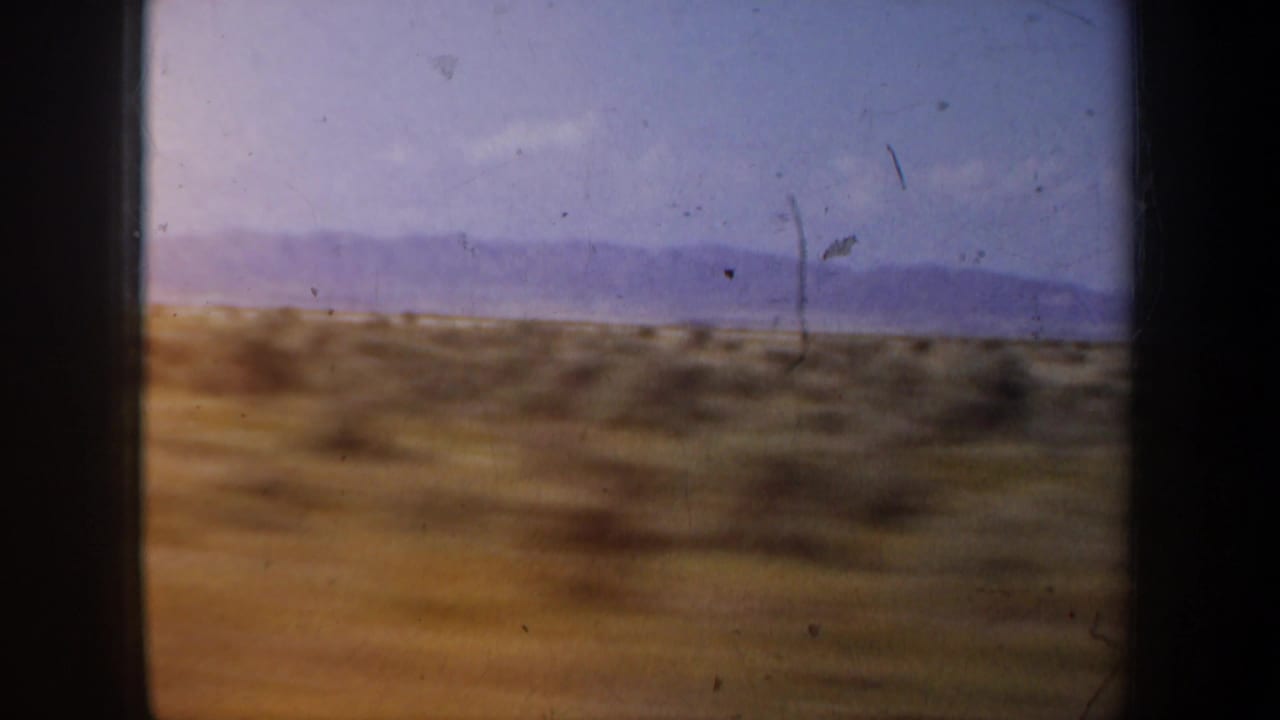 Scrub desert and distant hills seen through a dark vehicle-window frame.