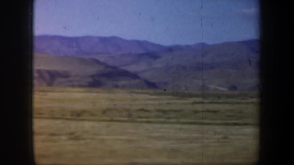 1957 Oregon 8mm travel frame showing open scrub plain, layered distant hills, pale sky, and vertical film marks from a moving vehicle.