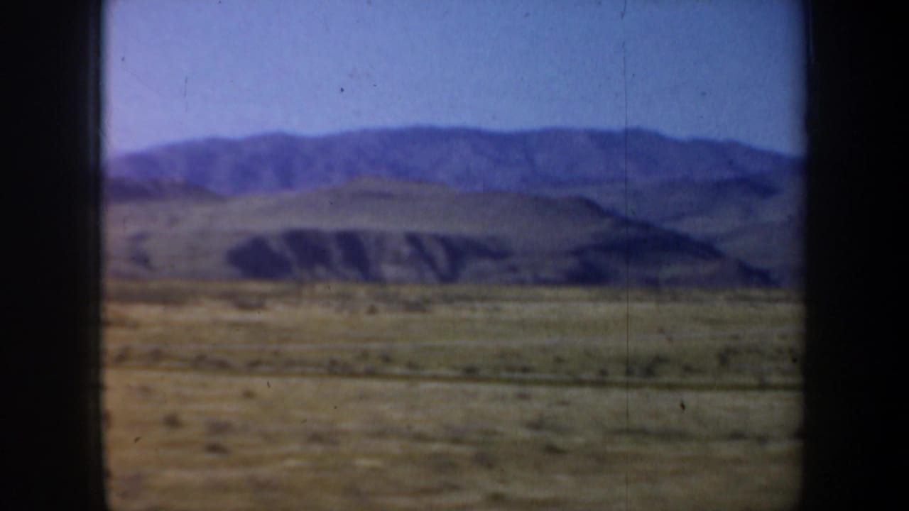Open arid plain with low hills and distant mountains seen through a vehicle window.