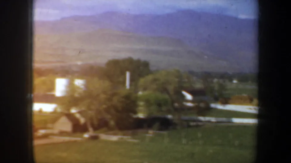 1957 Oregon home-movie view of a small rural settlement with fields, trees, buildings, and white silos below distant hills.
