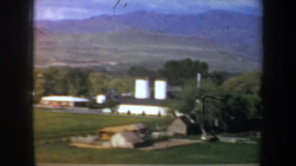 1957 Oregon frame showing a rural settlement with white silos, trees, low buildings, and mountain ridges in the background.