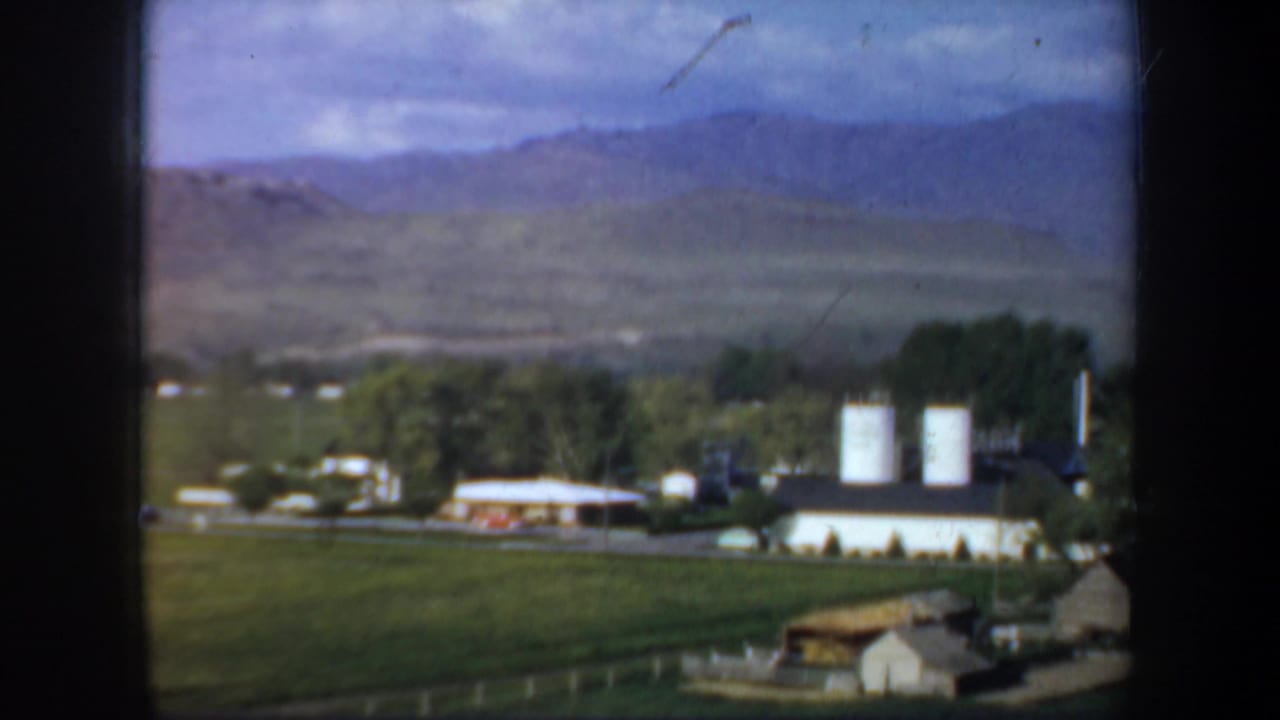 1957 Oregon 8mm frame showing white rural buildings, two silos, green fields, trees, and distant mountains from a moving viewpoint.