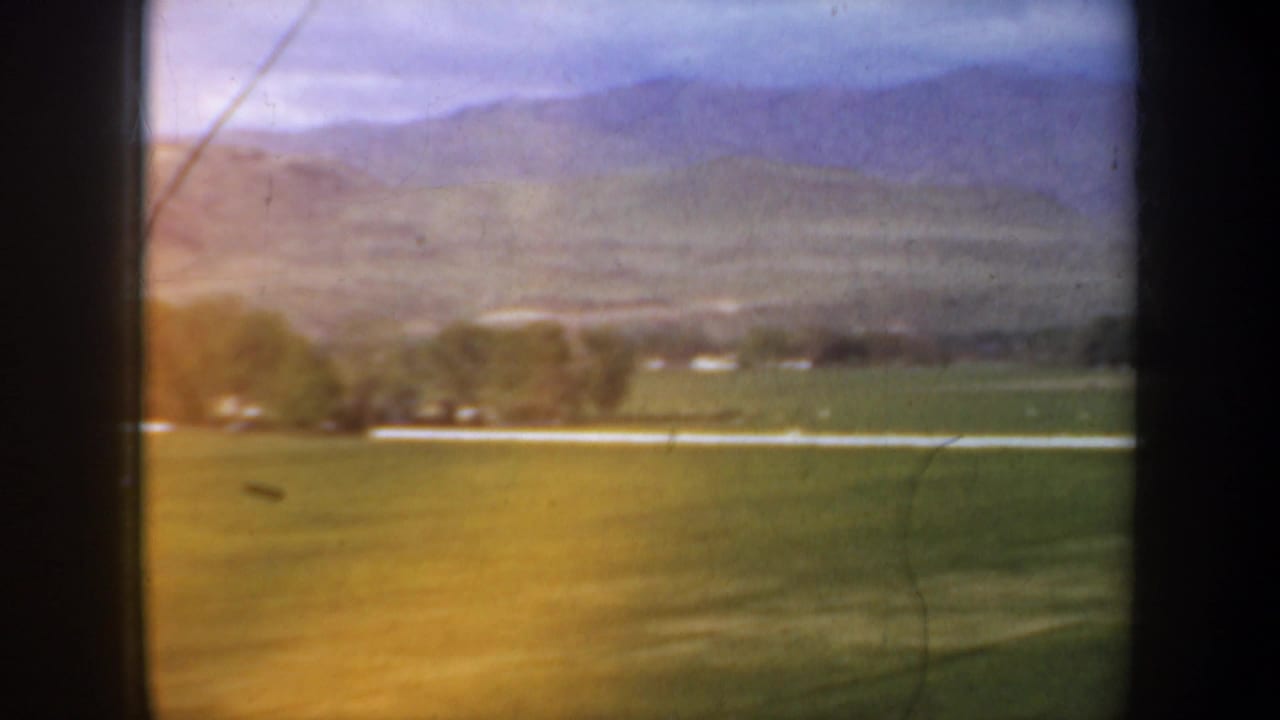 1957 Oregon roadside view across green fields toward low mountains, with a pale road line and trees near the horizon.