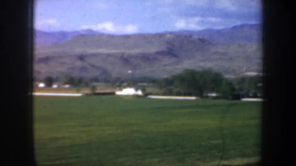 1957 Oregon frame with green fields, a light road or lane, trees, small pale buildings, and low mountains in the background.