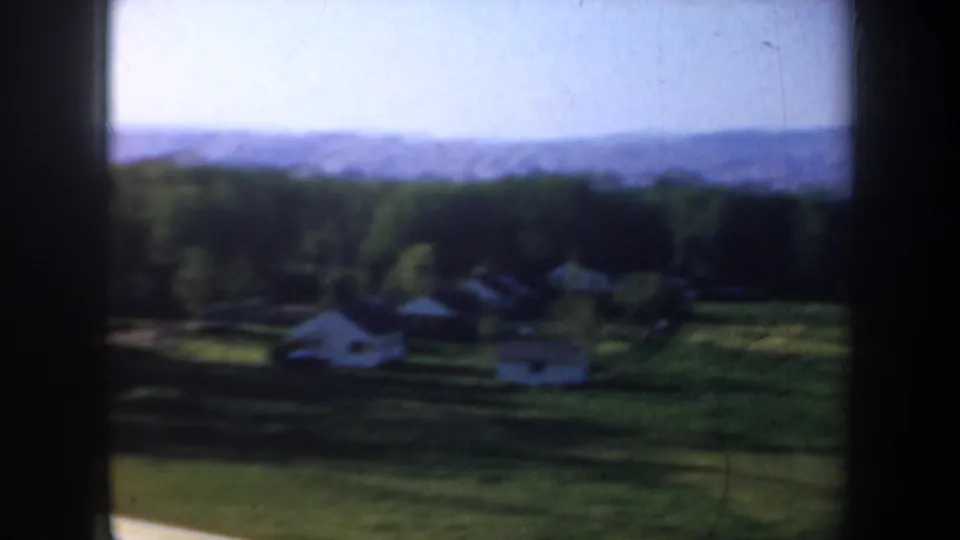 1957 Oregon home-movie frame looking over shaded rural buildings, trees, open land, and a hazy mountain horizon.