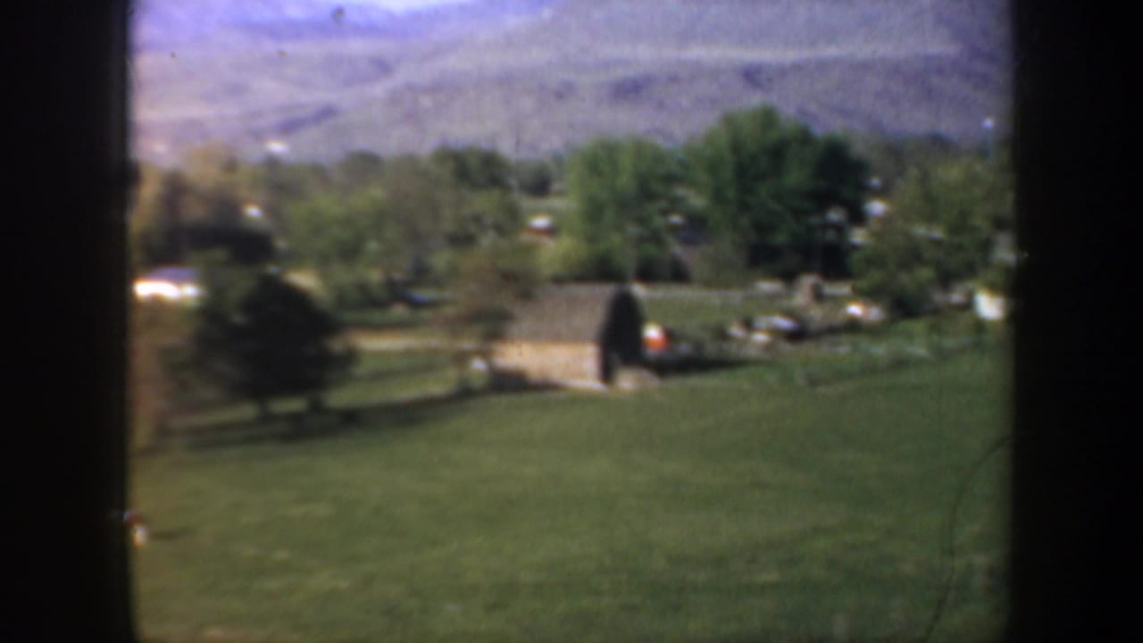 1957 Oregon home-movie frame showing green pasture, trees, small rural buildings, and mountains beyond a valley landscape.