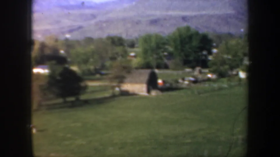 1957 Oregon home-movie frame showing green pasture, trees, small rural buildings, and mountains beyond a valley landscape.