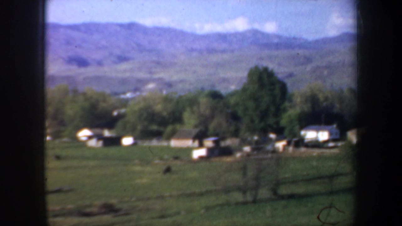 Open field with small buildings, trees, fencing, and mountains in the background.