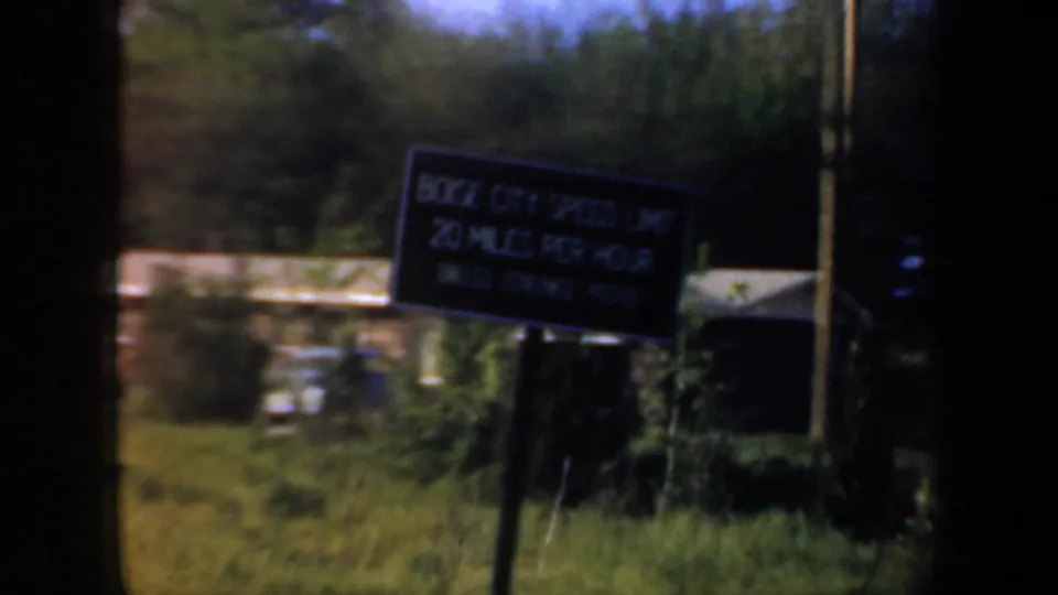 1957 Oregon roadside frame with a dark sign or billboard near grass, trees, a utility pole, and rural buildings in the background.