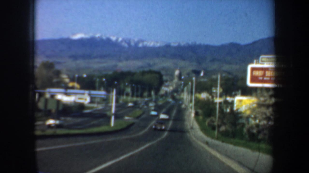 1957 home-movie road view from a car, showing light traffic, a roadside bank sign area, trees, and snow-capped mountains beyond the town.