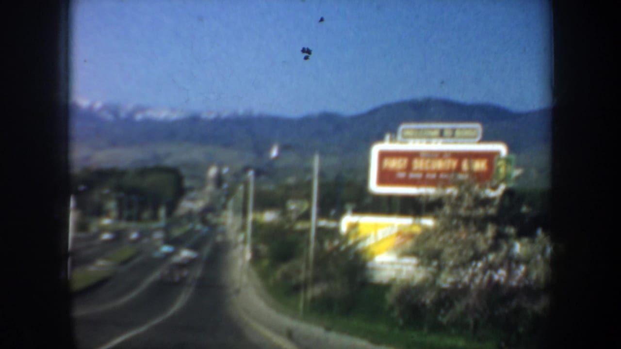 Cataloged Oregon 1957 road footage with cars on a broad street, a red First Security Bank sign, and mountains rising in the background.
